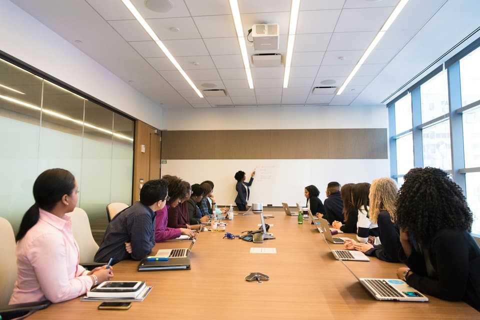 A working session in a boardroom: a dozen colleagues with laptops listen as one of them works through notes on a whiteboard.